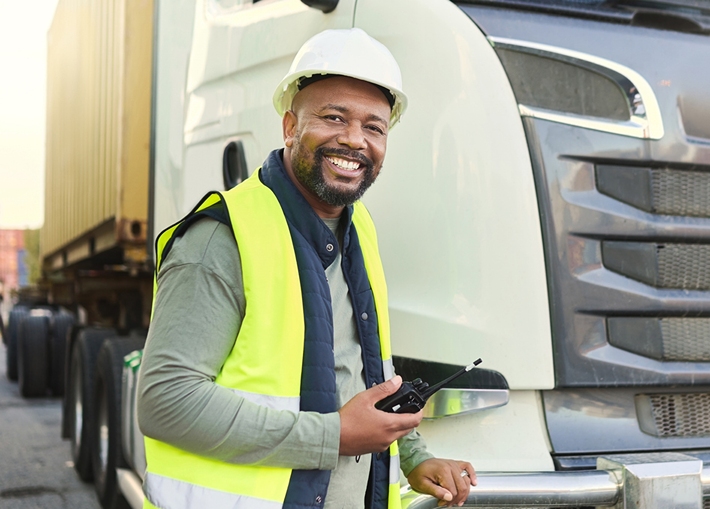 A smiling commercial truck driver wearing a high-visibility vest and a hard hat, holding a walkie-talkie, standing next to a large truck with a container in the background